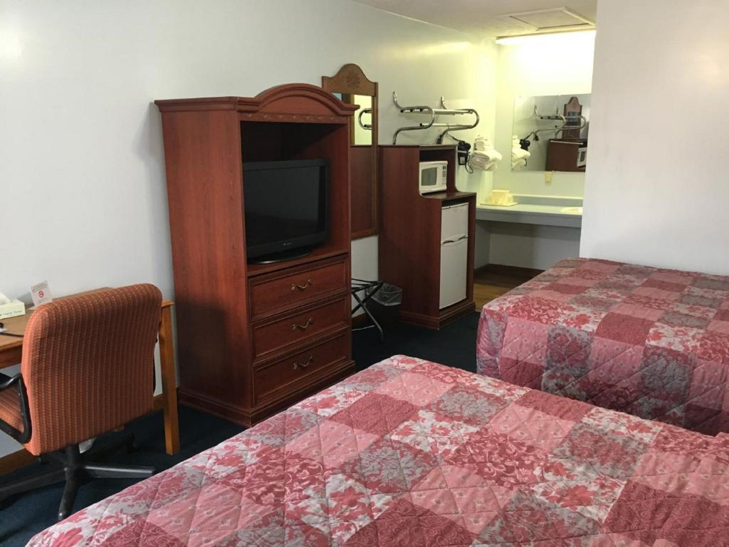 Habitación de hotel con dos camas, un aparador de madera con televisor, un escritorio con silla y un baño con espejo y lavamanos.