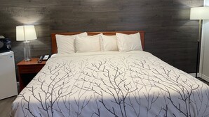 Une chambre d’hôtel avec une tête de lit en bois foncé, un couvre-lit blanc orné d’un motif de branches d’arbres, une lampe de chevet et une petite table de chevet blanche avec un téléphone.