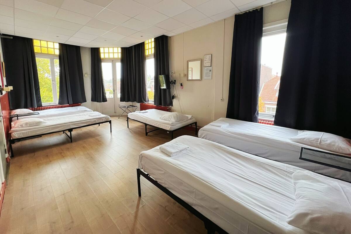 Une chambre d’hôtel avec trois lits simples, un plancher en bois, une fenêtre avec des rideaux et une vue sur l’extérieur.