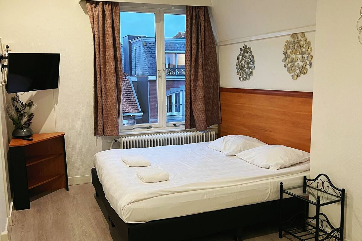Une chambre à coucher bien rangée, avec un lit, une table de chevet sur laquelle se trouve un téléviseur à écran plat, un radiateur et une fenêtre avec des rideaux.