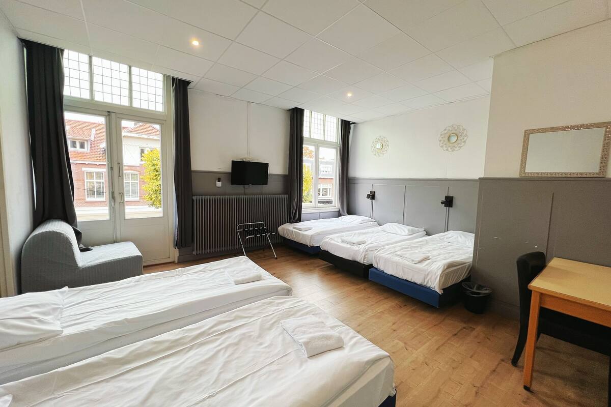 Une chambre d’hôtel avec deux lits, un bureau, une chaise, une télévision et de grandes fenêtres avec des rideaux.