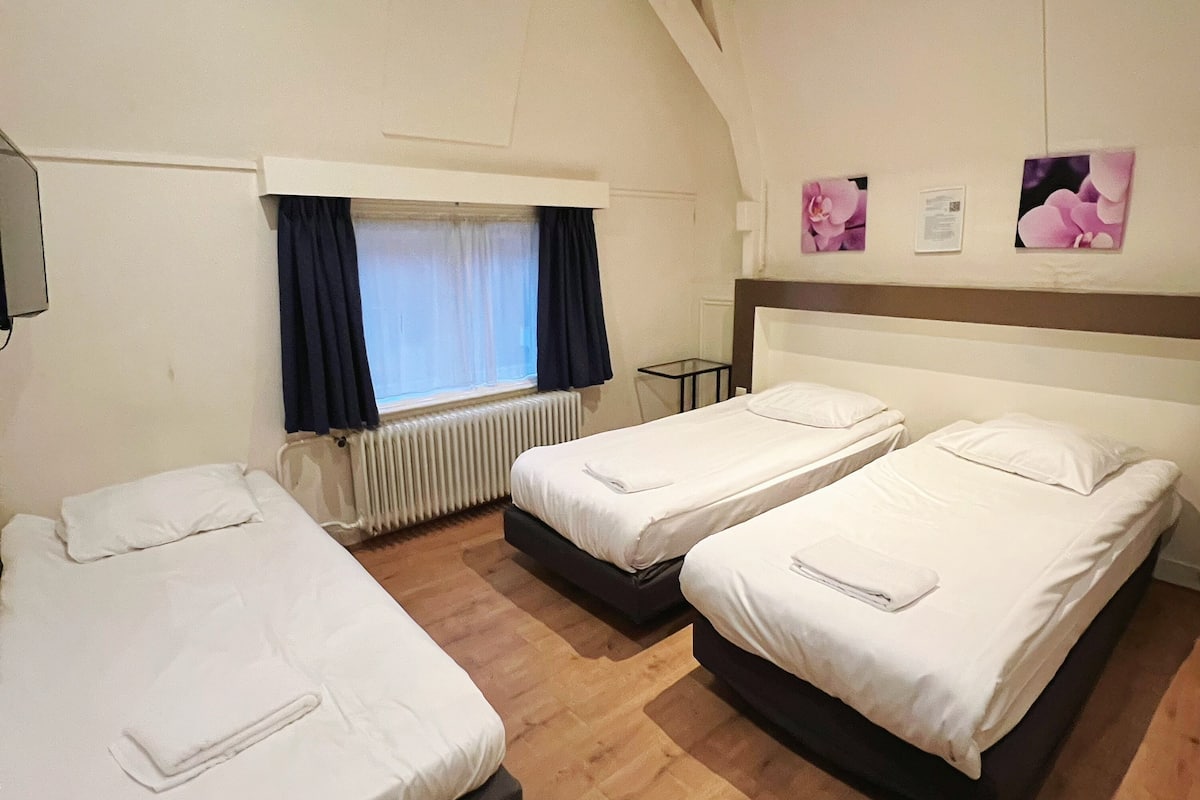 Une chambre d’hôtel avec trois lits simples, une fenêtre avec des rideaux, un radiateur et une petite table.