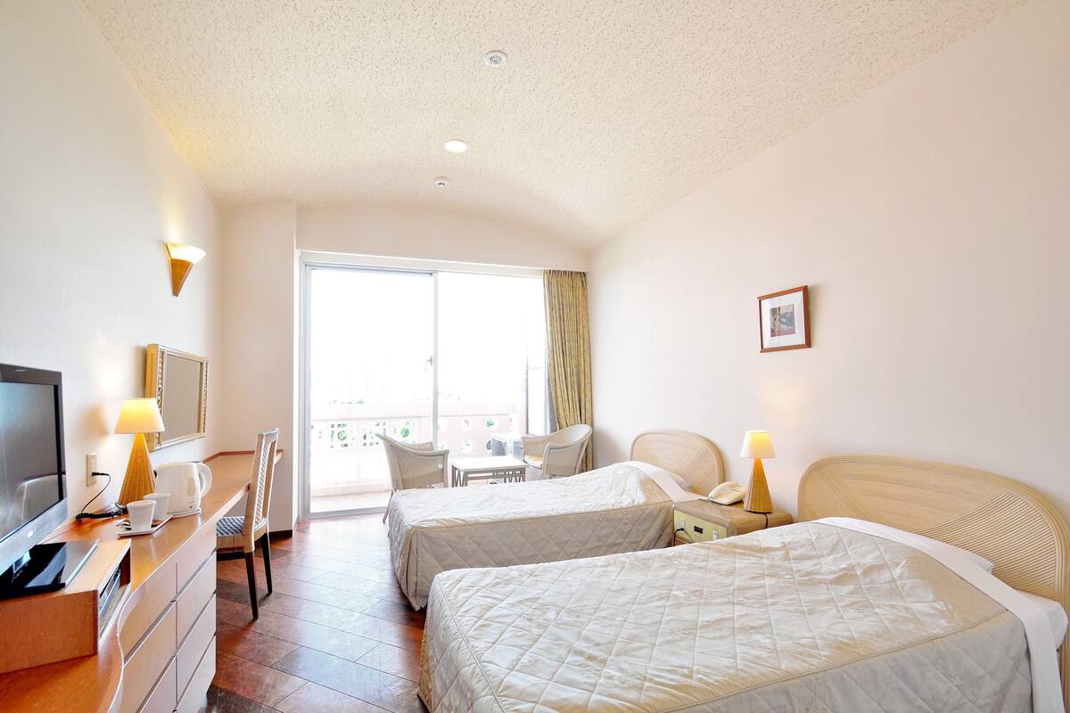 Hotellihuone, jossa on kaksi sänkyä, työpöytä, tuoli, televisio ja parveke, jolta on näkymä.