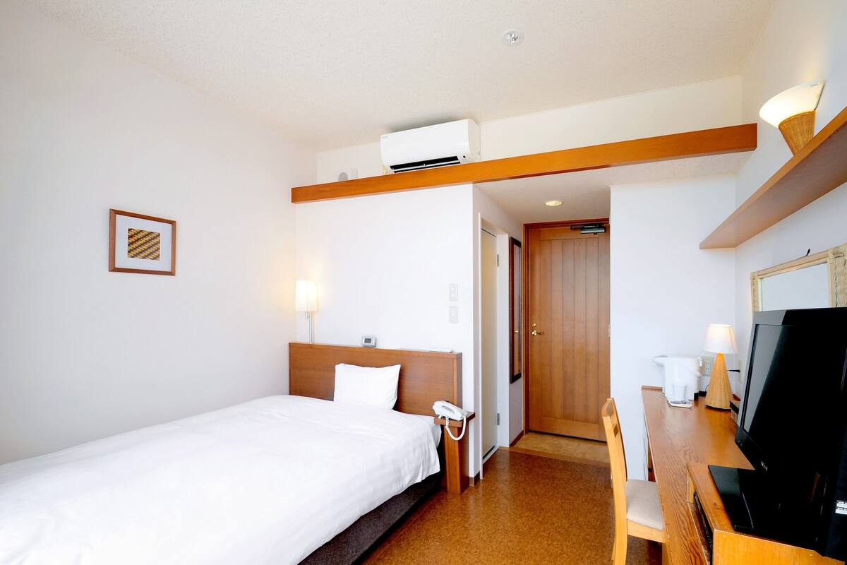 Hotellihuone, jossa on sänky, yöpöytä, televisio ja työpöytä tuolilla.
