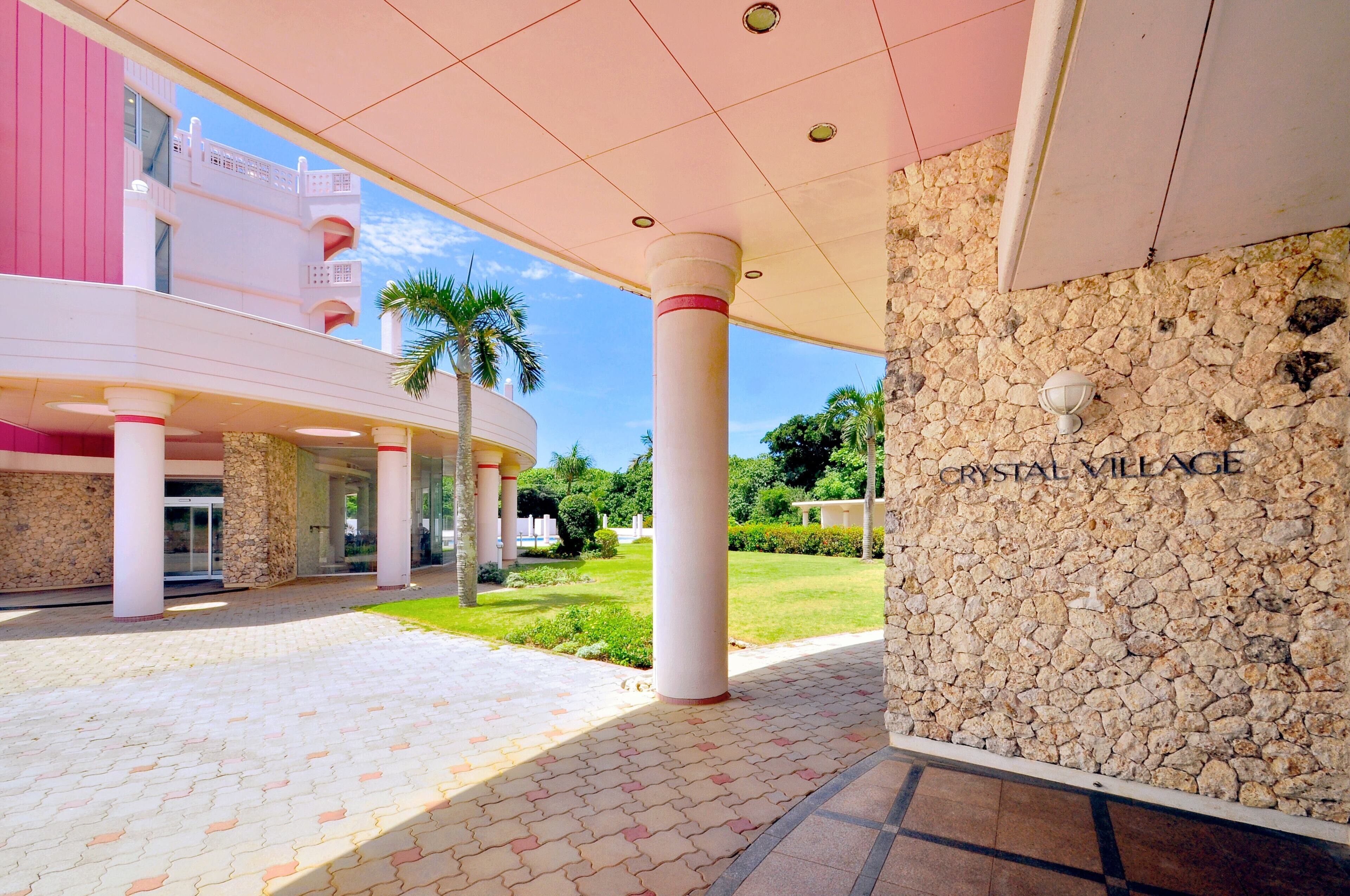 A building entrance with a stone wall labeled "CRYSTAL VILLAGE," a covered walkway, and a palm tree in the background.