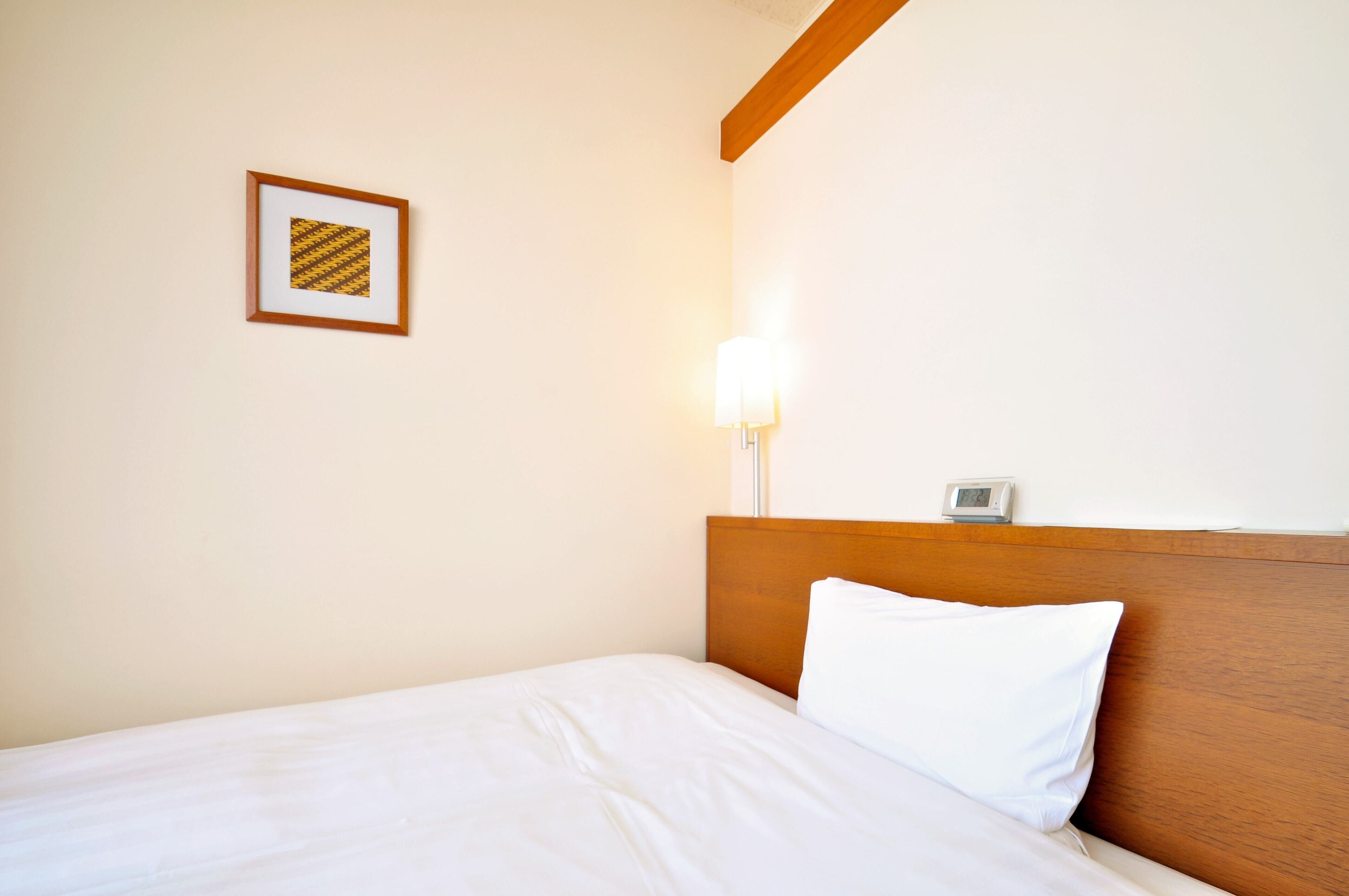 A hotel room with a wooden headboard, a white bed, a framed picture on the wall, and a bedside lamp.