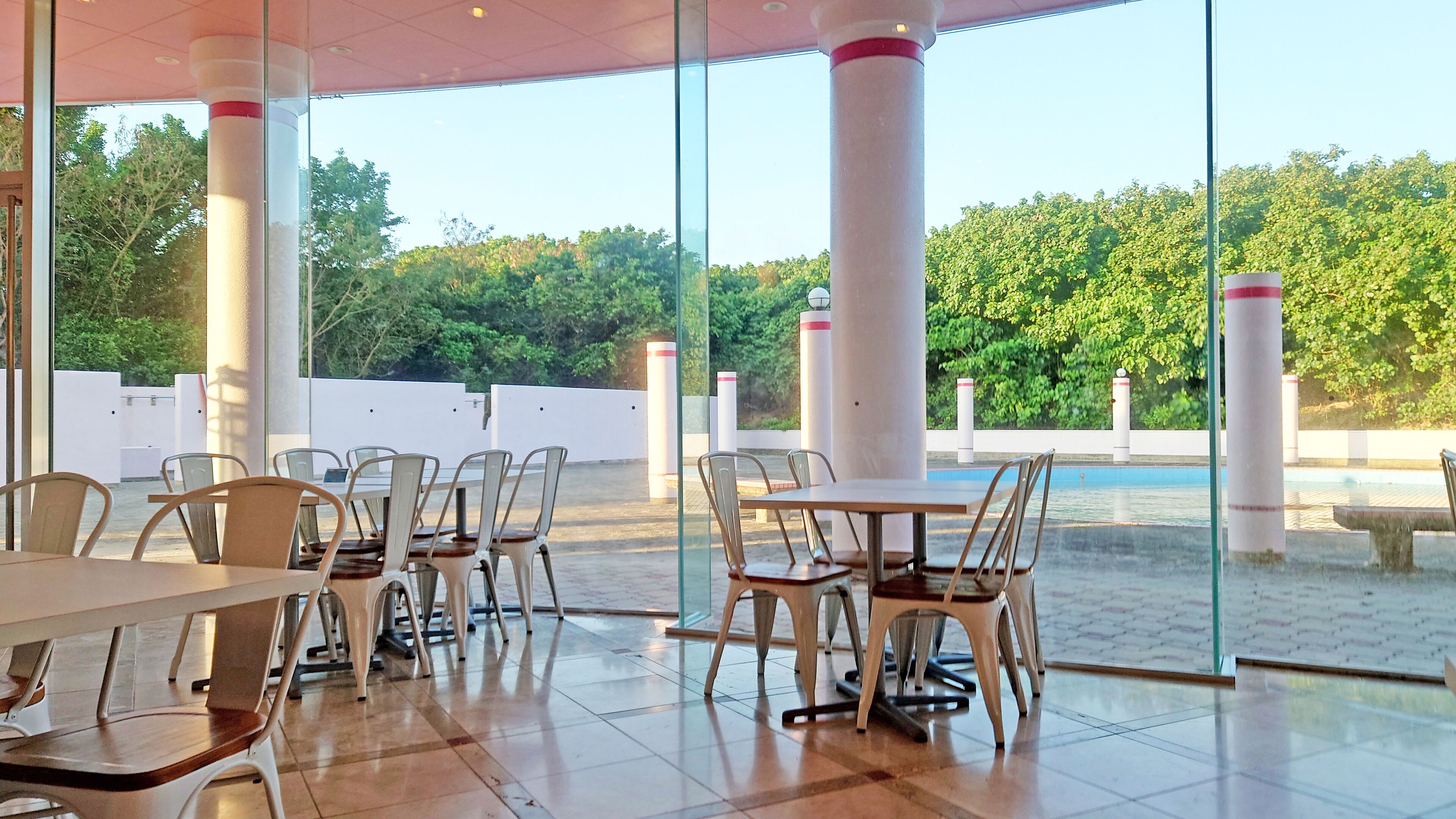 Un espacio de comedor al aire libre con mesas y sillas, amplios ventanales y vistas a zonas verdes y una piscina.