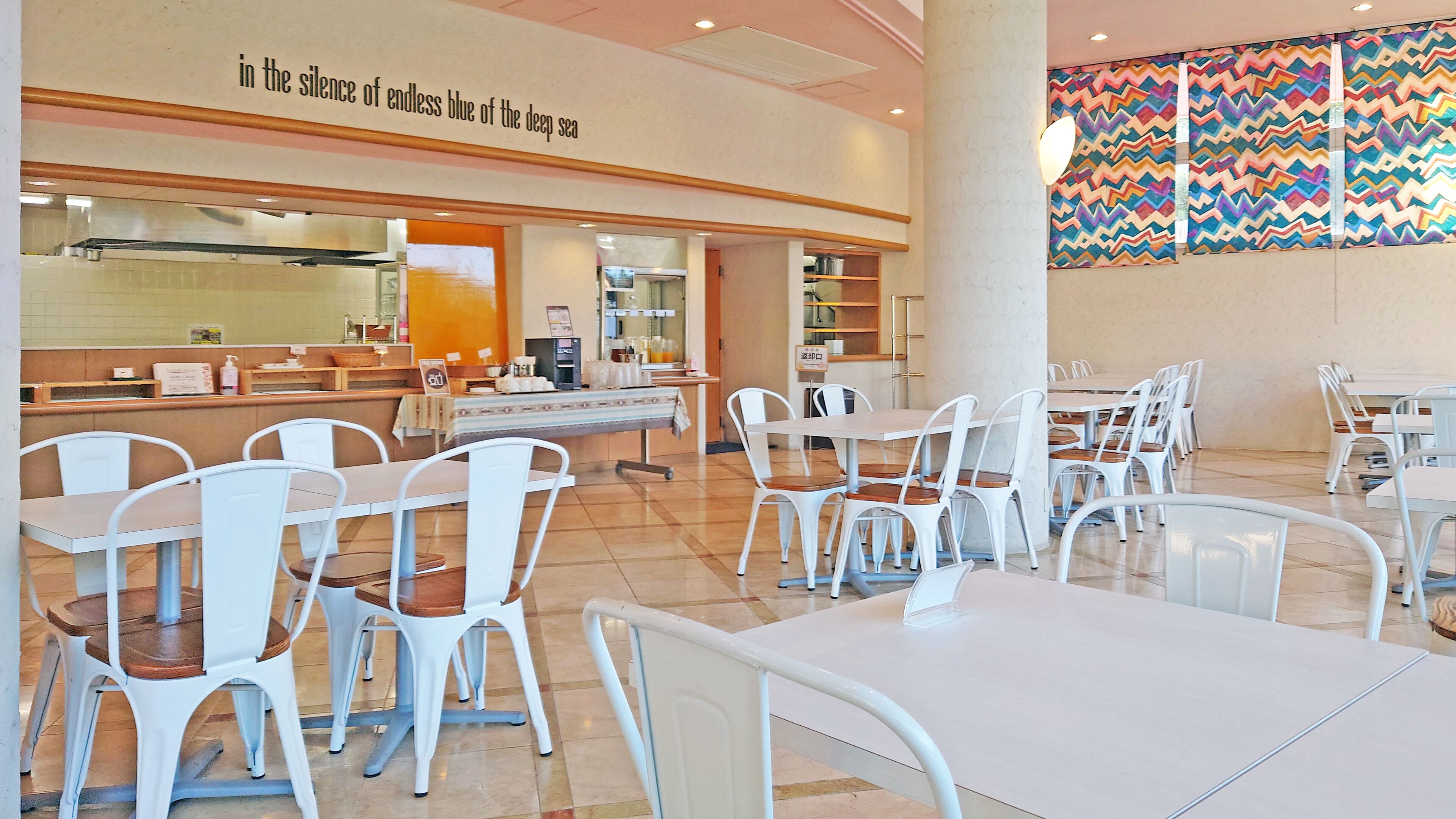 A spacious restaurant with white chairs and tables, a counter with a menu, and a colorful wall mural.
