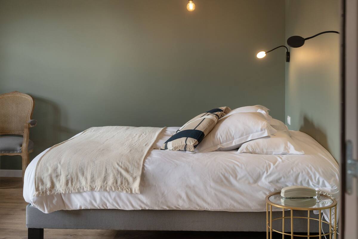 A neatly made bed with white linens and a beige throw blanket, a wicker chair, and a small round table with a lamp.