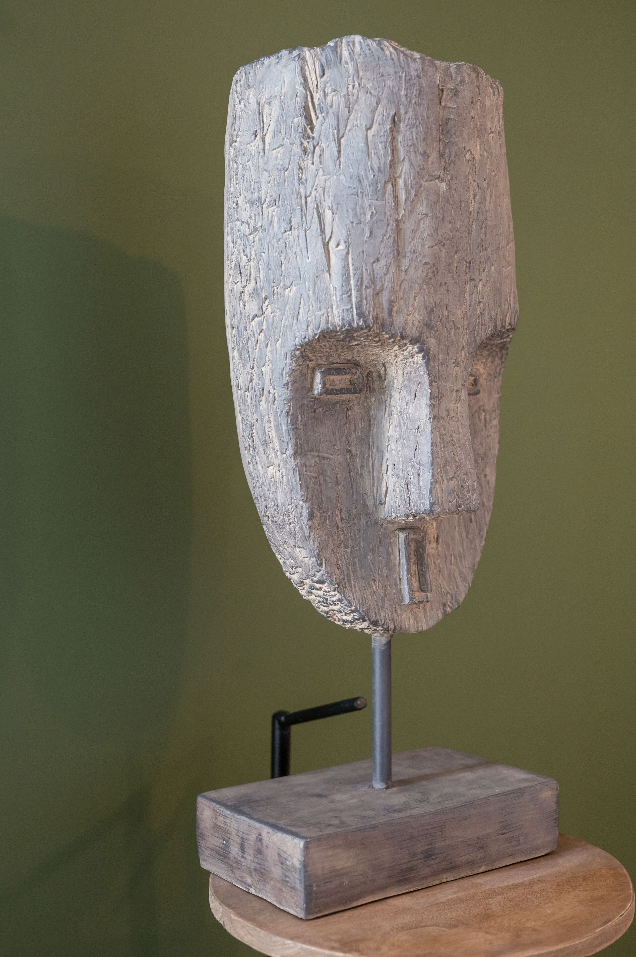 A sculpture of a face mounted on a wooden base, displayed against a green wall.
