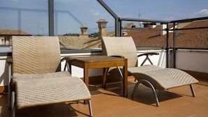 Eine Terrasse mit zwei Korbstühlen und einem kleinen Holztisch mit Blick auf eine Stadtlandschaft und traditionelle Dächer.
