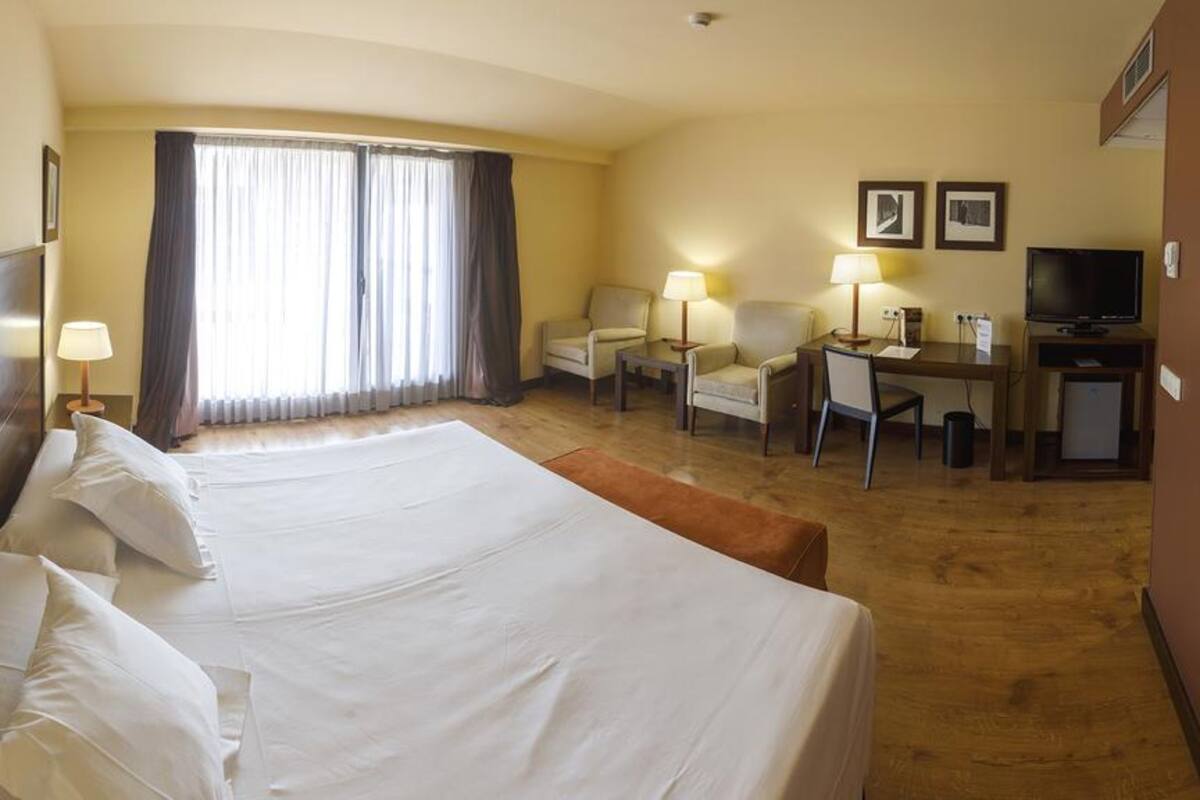Ein Hotelzimmer mit einem großen Bett, zwei Sesseln, einem Schreibtisch, einem Fernseher und einem Fenster mit Vorhängen.
