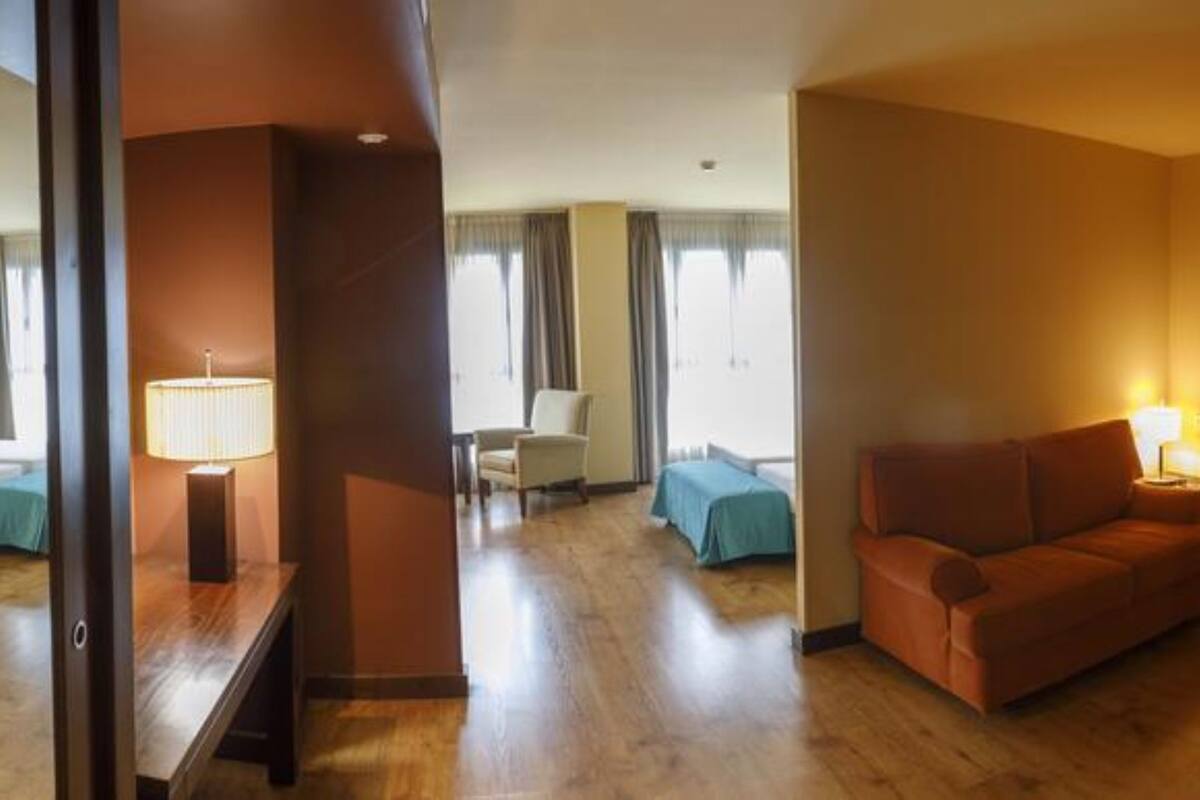 Ein Hotelzimmer mit zwei orangenen Sofas, einem Holztisch, einem Bett mit blauer Bettdecke und einem Fenster mit Vorhängen.