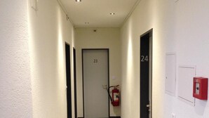 Kapea käytävä, jossa on ovet numeroilla 23 ja 24, seinälle asennettu palosammutin ja kattoon upotetut valaisimet.
