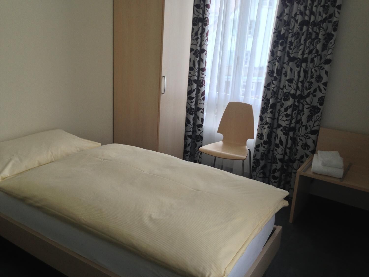 Una habitación sencilla y limpia con una cama, una silla y una pequeña mesa con una toalla.