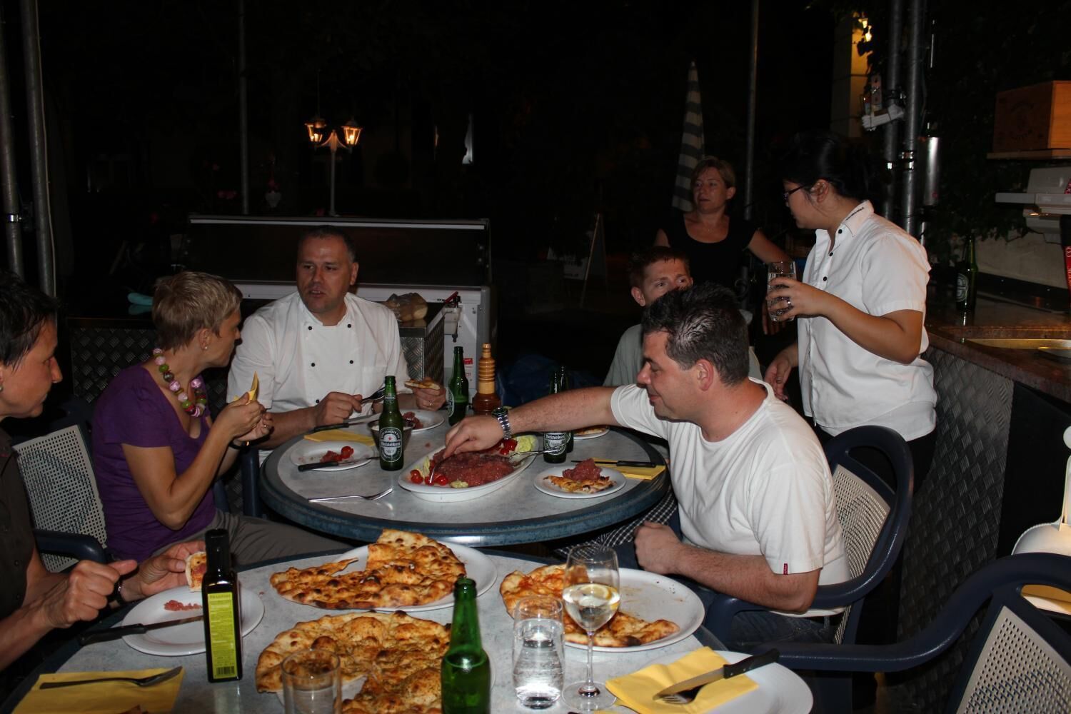 Un grupo de personas cenando en un restaurante al aire libre con mesas y sillas redondas, en un ambiente oscuro y tenue.