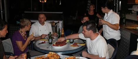 Un grupo de personas cenando en un restaurante al aire libre con mesas y sillas redondas, en un ambiente oscuro y tenue.