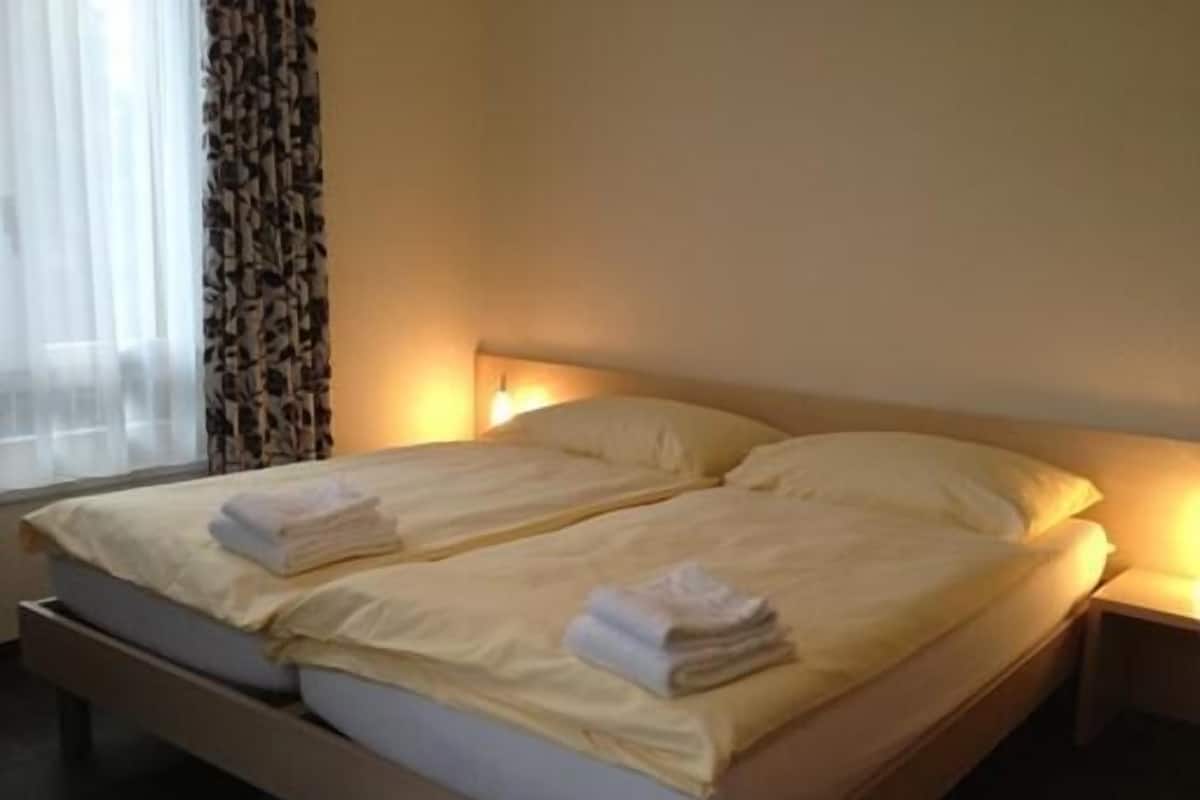 A double bed with two pillows and folded towels, a bedside table, and a window with curtains.