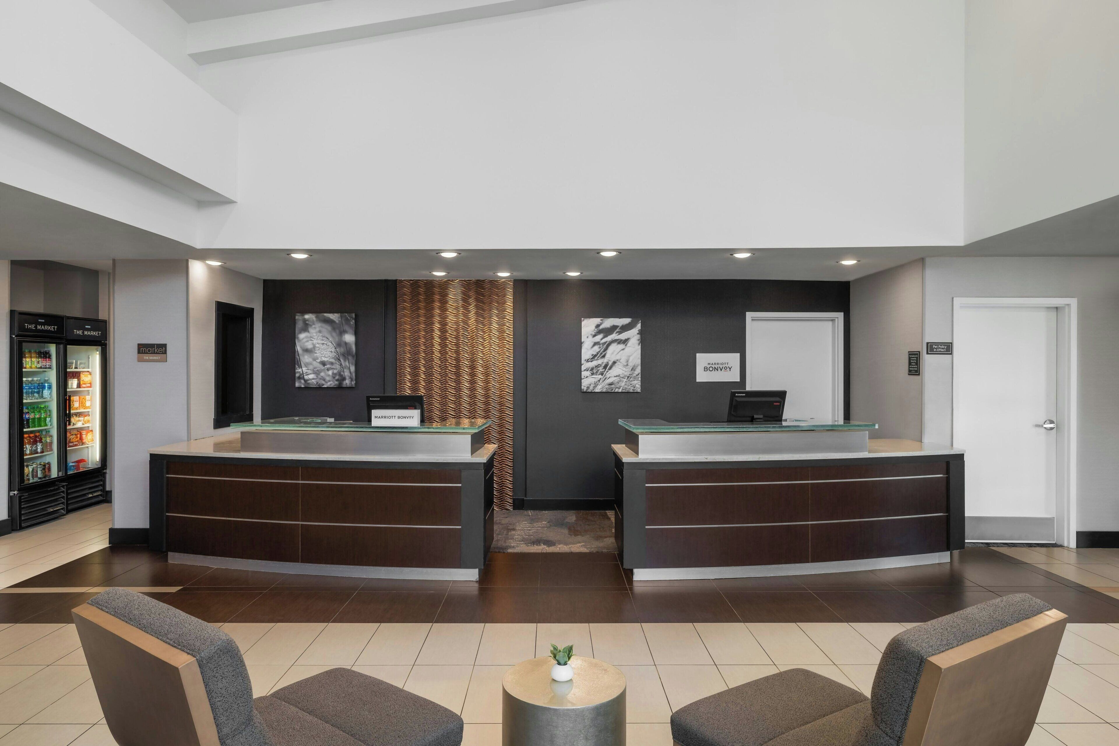 A hotel reception area with two reception desks, a seating area with chairs, a small table with a plant, and a refrigerator stocked with beverages.