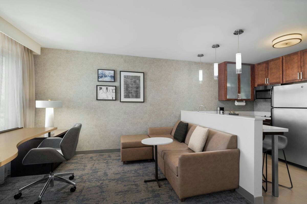 A modern hotel room with a kitchenette, a sofa, a small round table, and a desk with a chair.