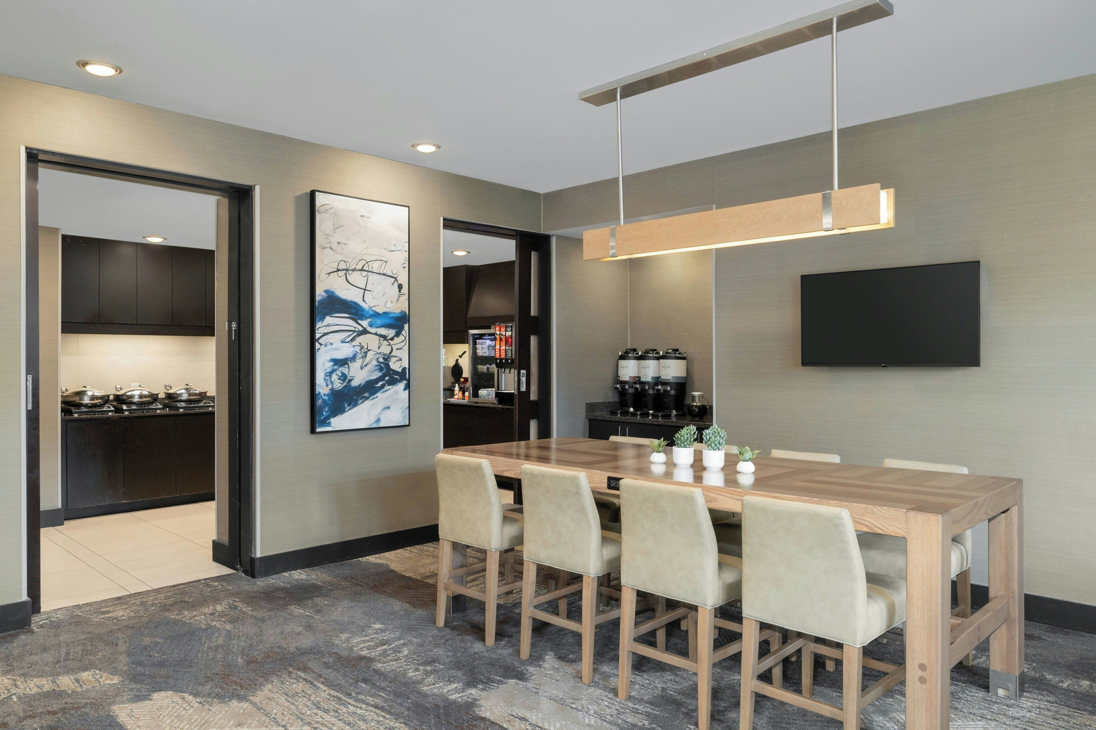 Un espace repas moderne avec une table et des chaises en bois, un téléviseur à écran plat fixé au mur et une cuisine visible à travers une porte ouverte.