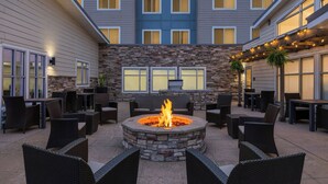 An outdoor patio area with a stone fire pit surrounded by wicker furniture, a stone wall, and a building with multiple windows in the background. - Residence Inn Champaign (Champaign)