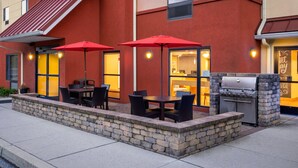 An outdoor patio with tables and chairs, a grill, and red umbrellas. - TownePlace Suites York (York)