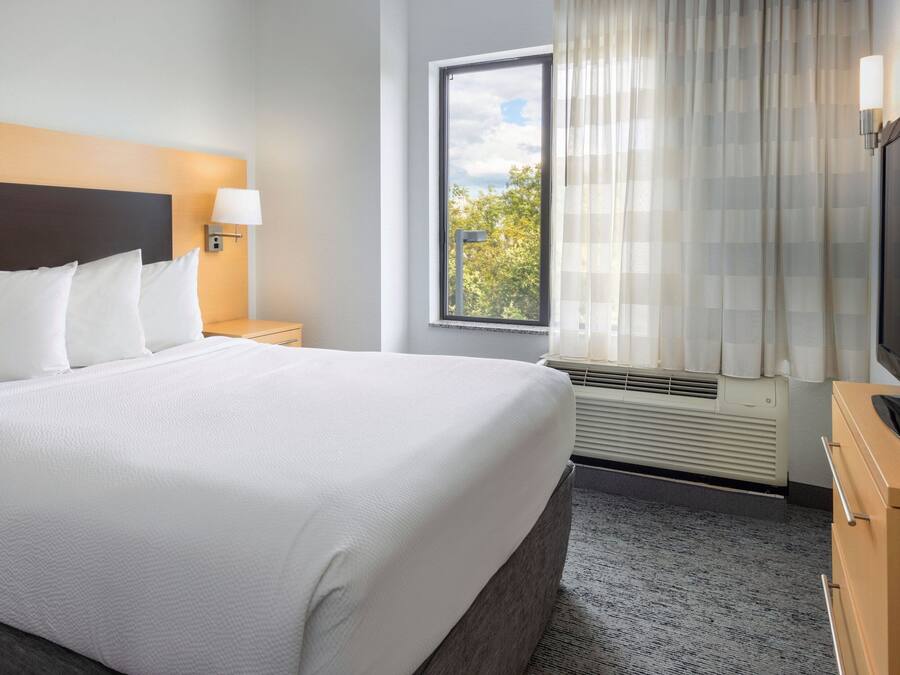 A hotel room with a large bed, a TV, a window with curtains, and a nightstand with a lamp.