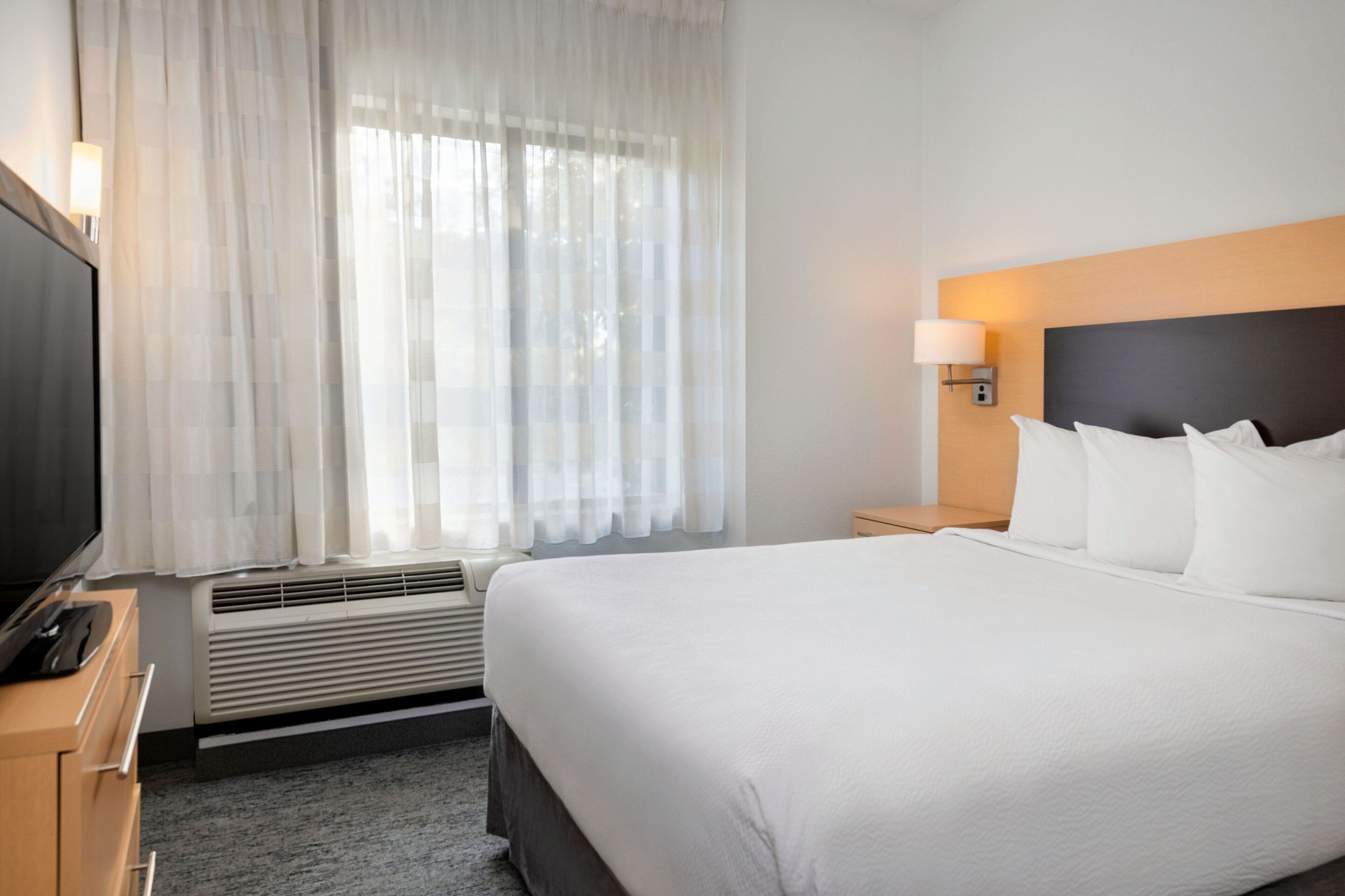 A hotel room with a bed, a TV on a wooden cabinet, a heater, and a window with sheer curtains.