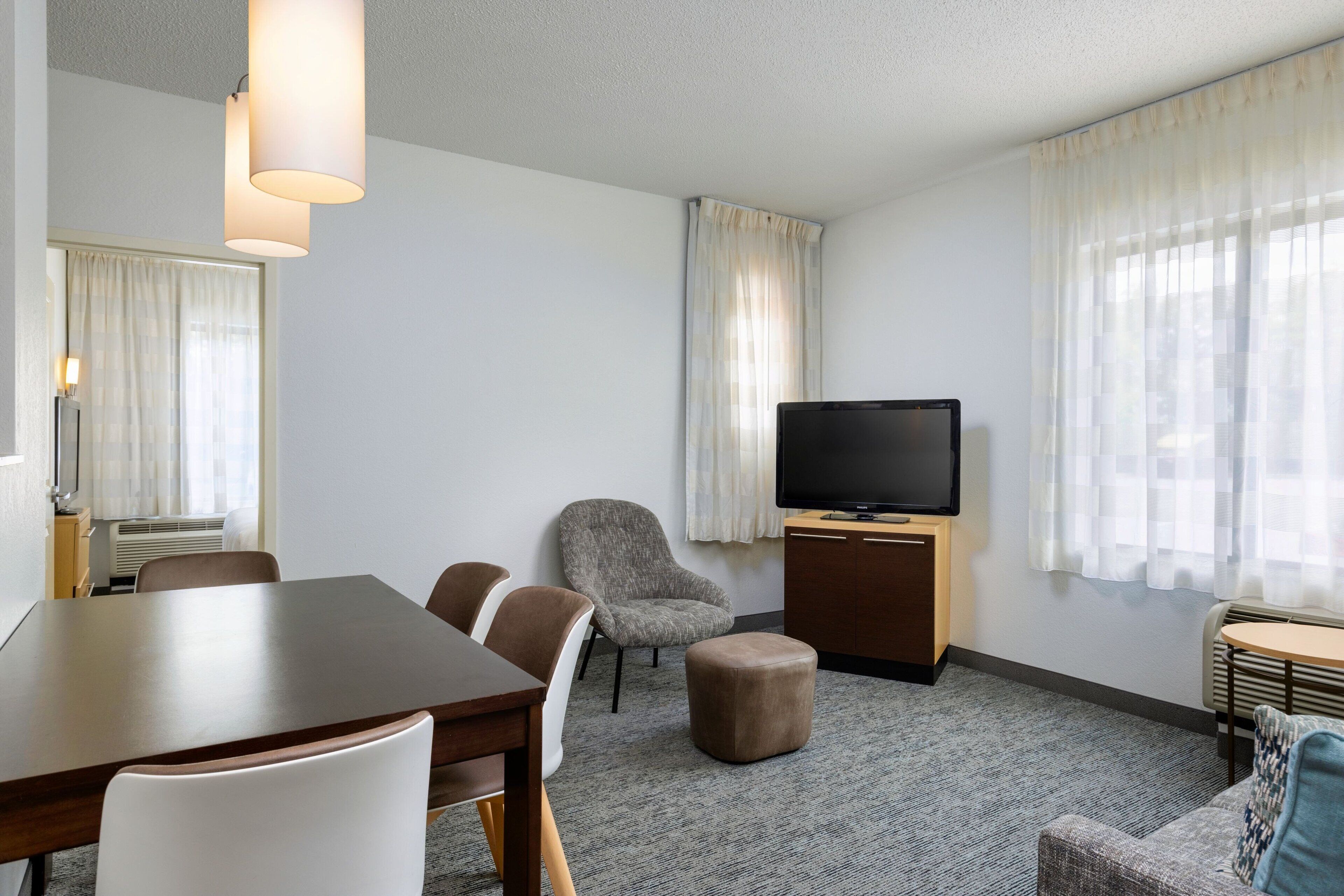 A hotel room with a dining table, chairs, a sofa, a television, and a small table with a lamp.