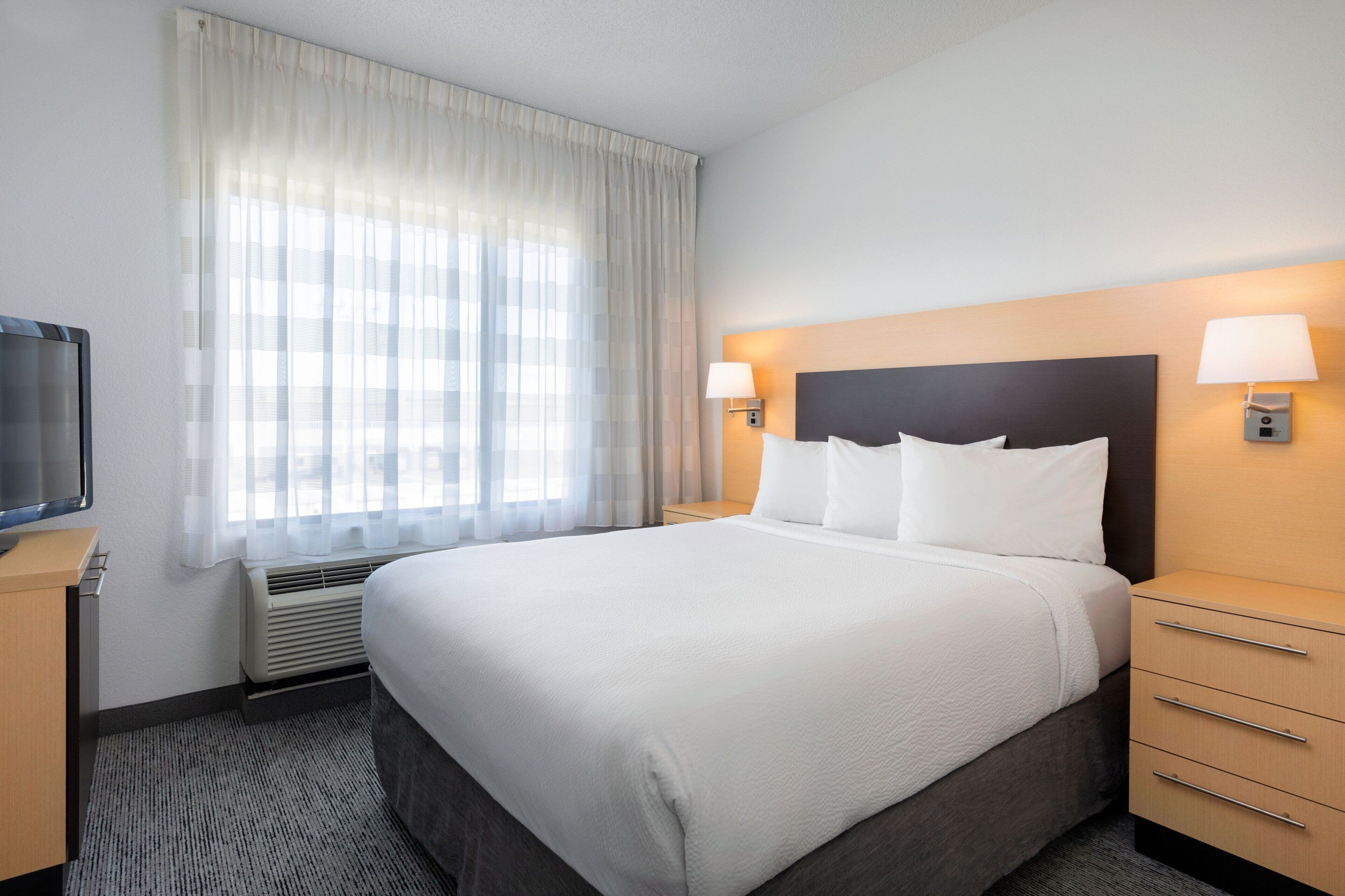 A hotel room with a large bed, two bedside lamps, a flat-screen TV, and a window with sheer curtains.
