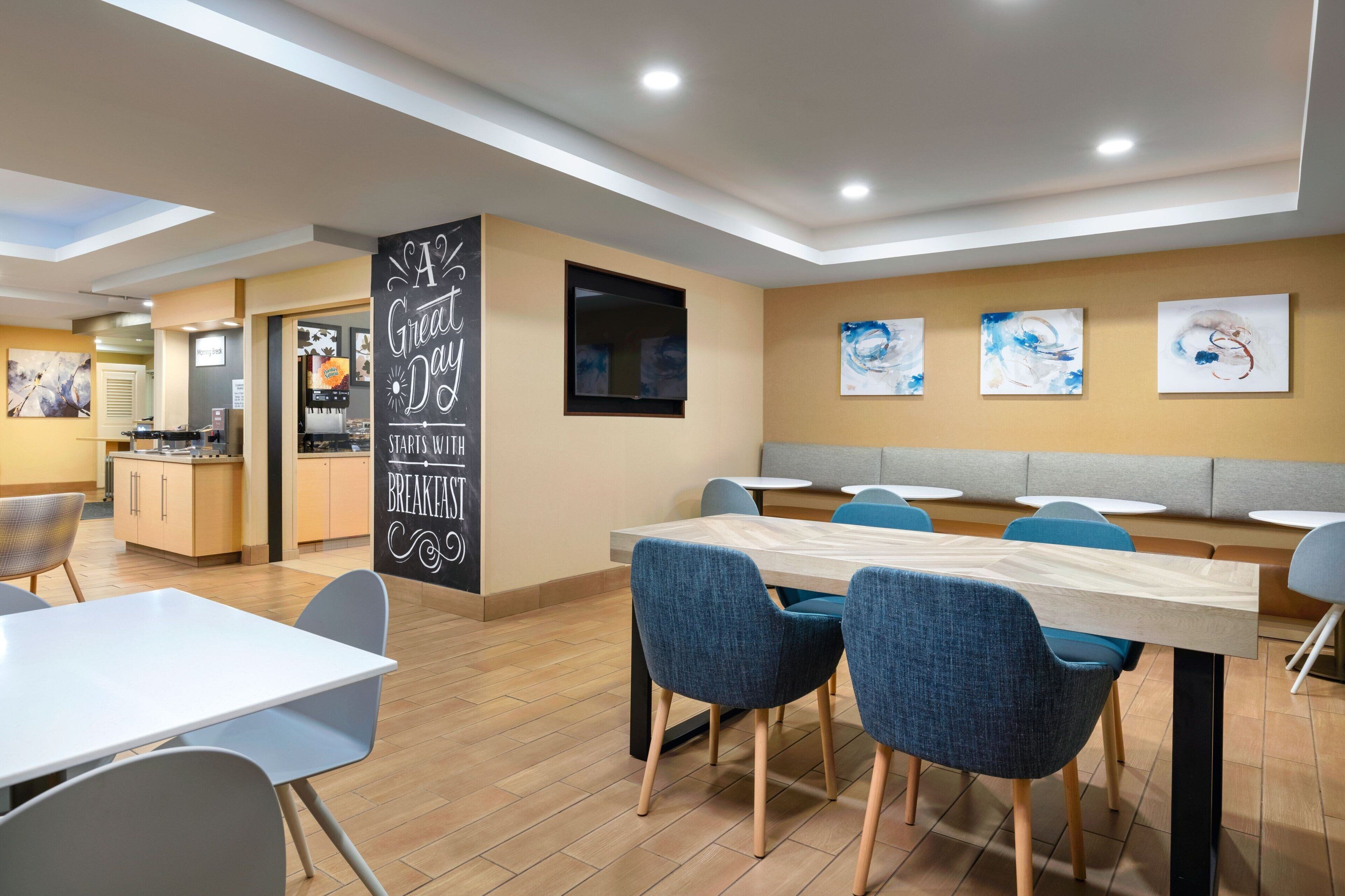 A modern dining area with a long table and blue chairs, a kitchen in the background, and abstract art on the walls.