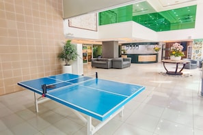 A ping pong table in a hotel lobby with a reception desk, seating area, and a green-tiled ceiling.