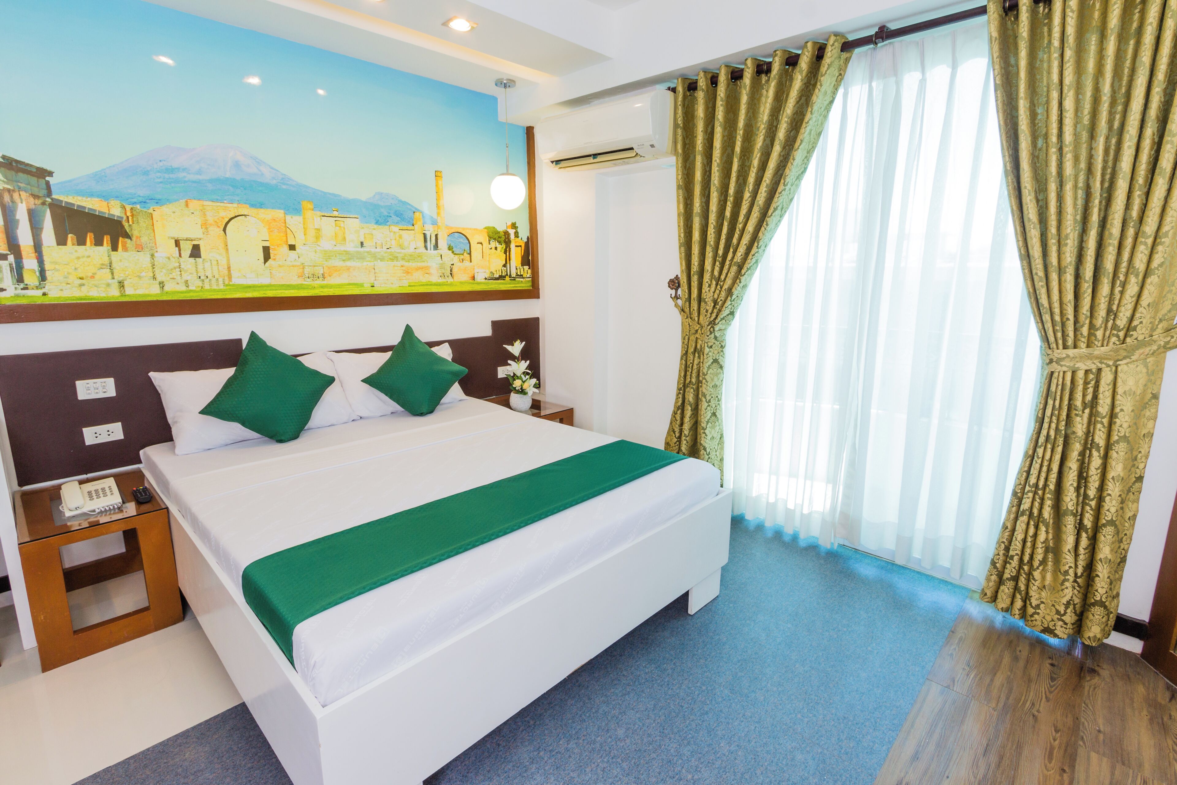 A hotel room with a bed, green pillows, a nightstand with a phone, a painting on the wall, and a window with curtains.
