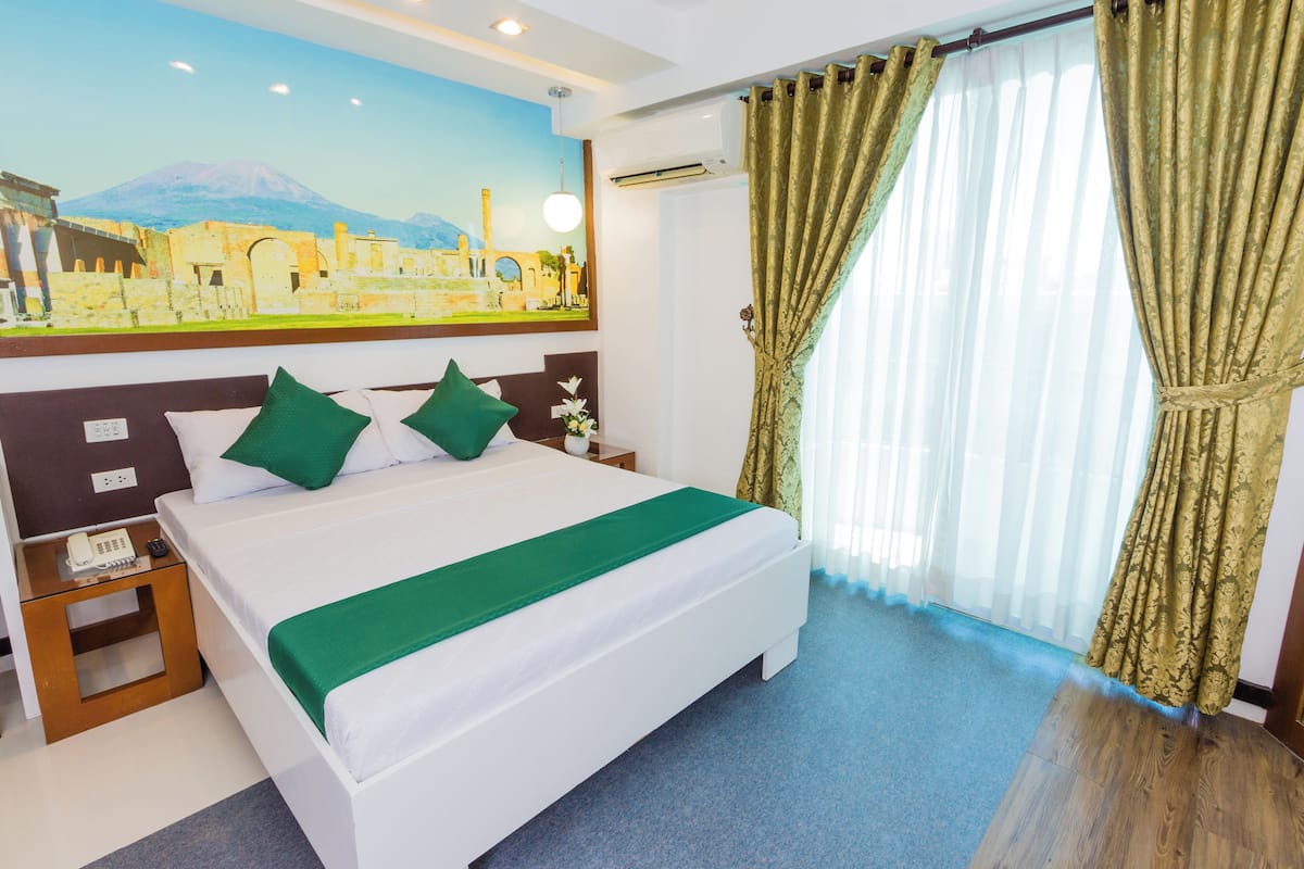 A hotel room with a bed, green pillows, a nightstand with a phone, a painting on the wall, and a window with curtains.