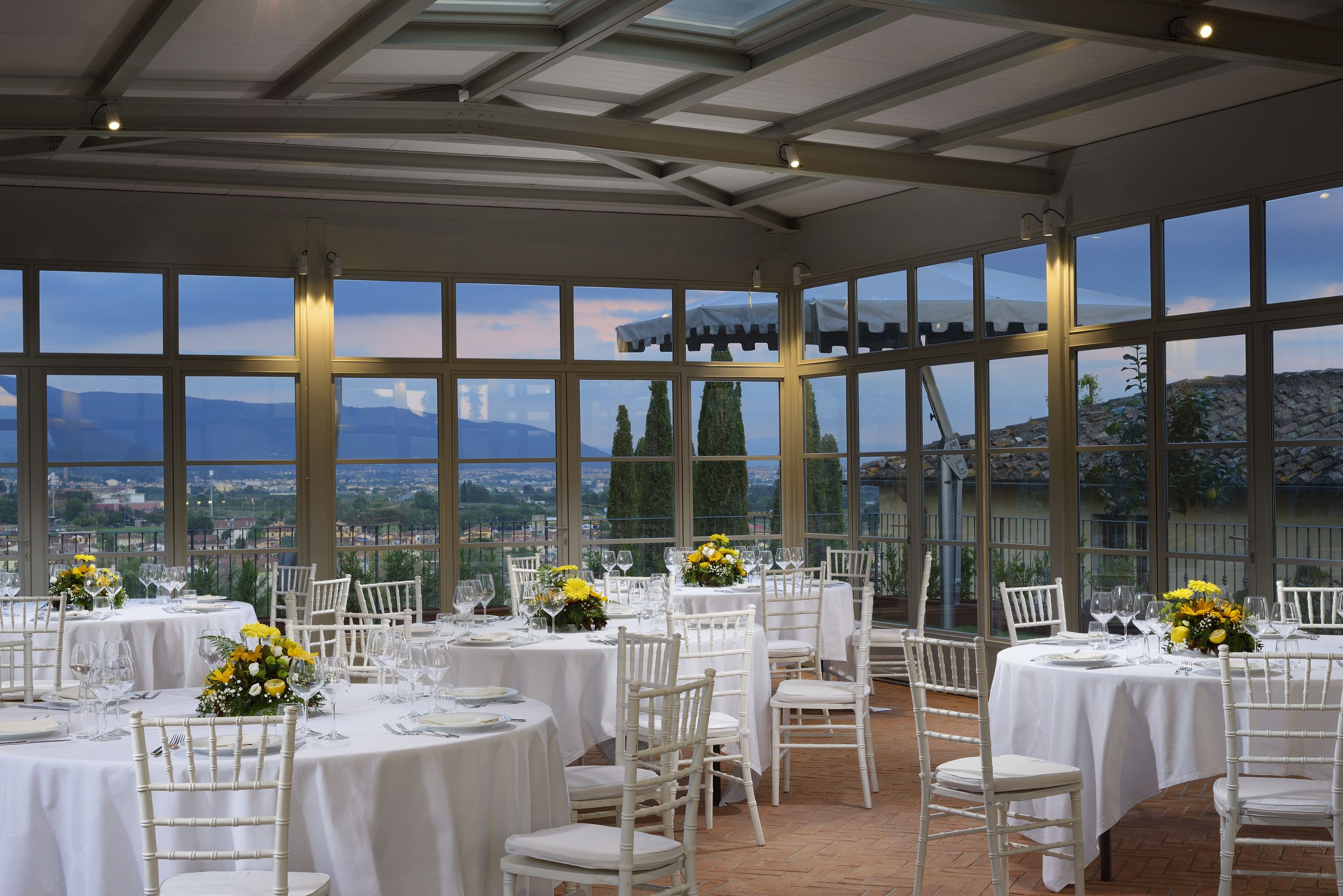 A dining area with round tables set for a meal, large windows with a view of the landscape, and white chairs.