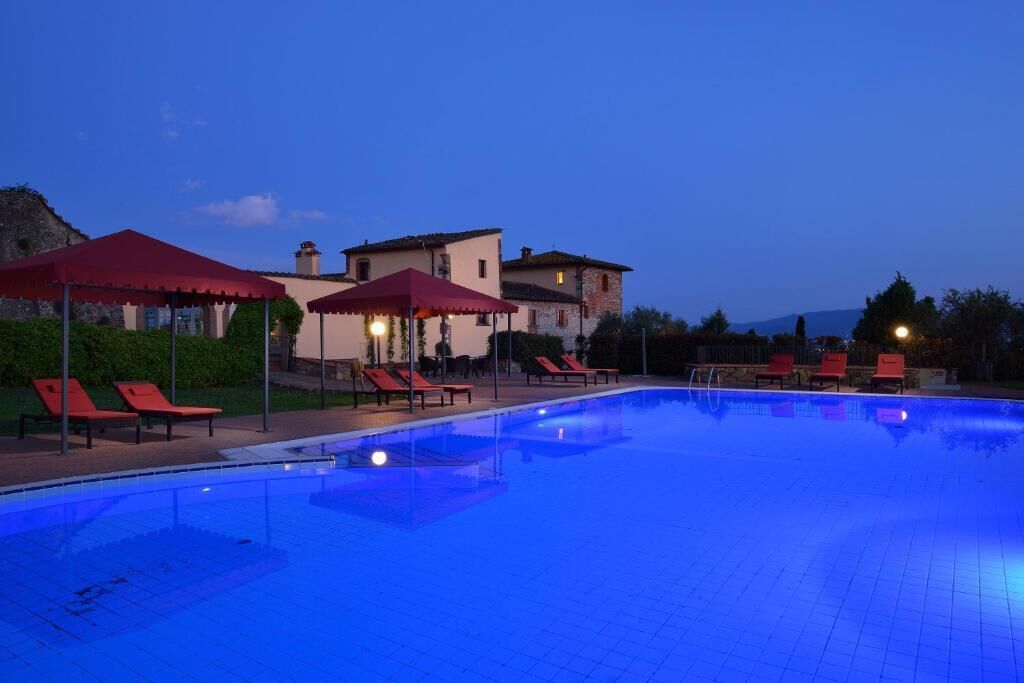 A swimming pool with red umbrellas and lounge chairs, surrounded by buildings with a mountainous backdrop.