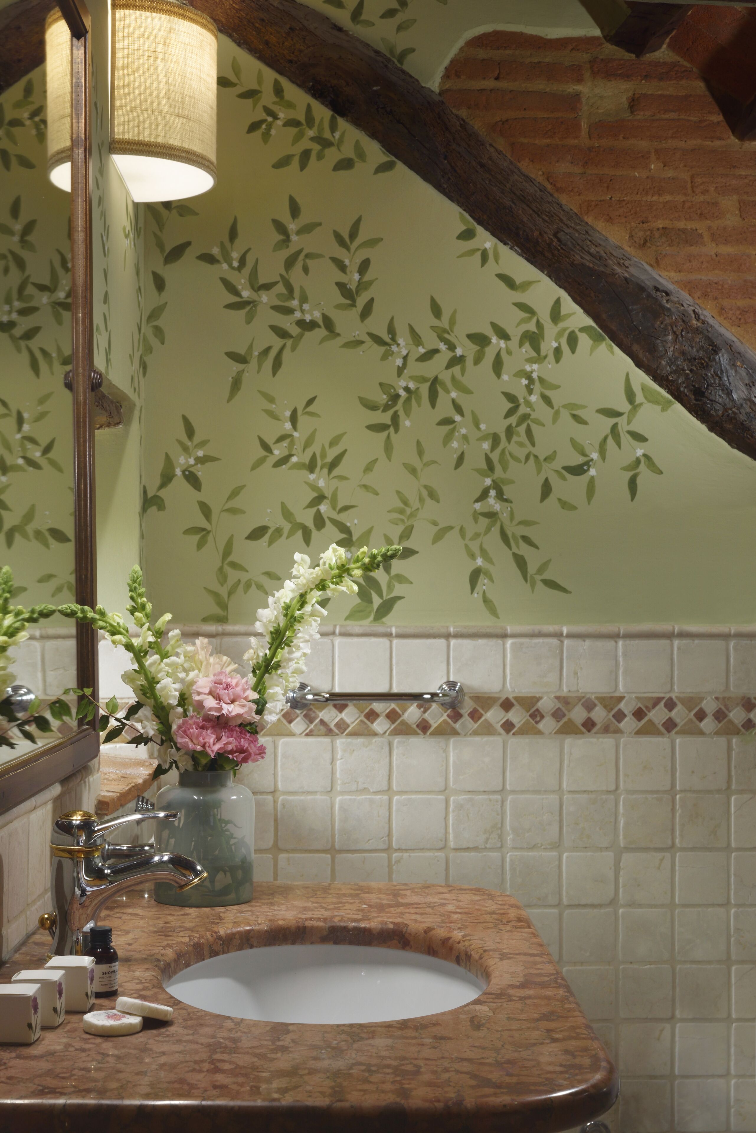 A bathroom with a marble sink, a mirror, a vase with flowers, and a wall with a leafy pattern.