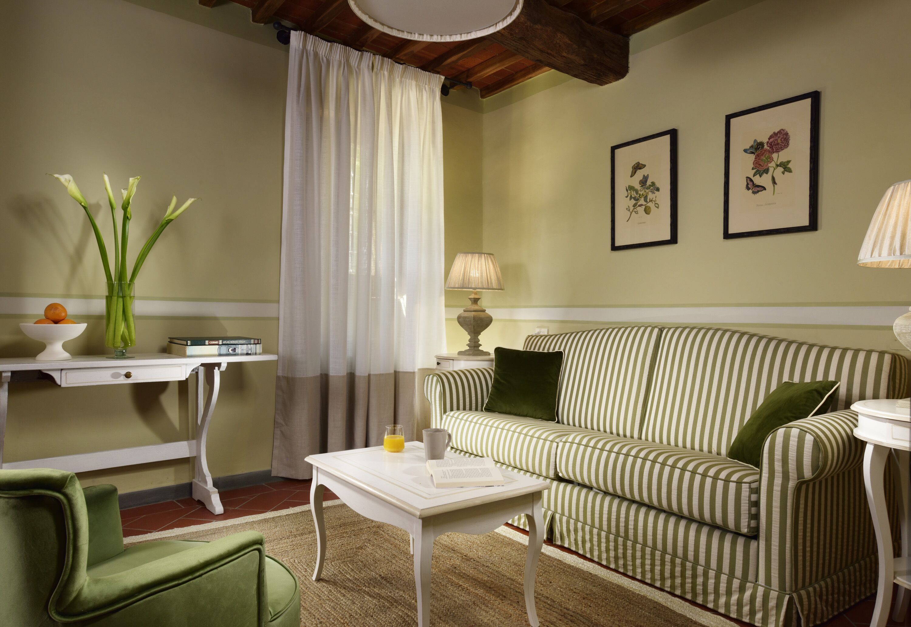 A cozy living room with a striped sofa, a small white table with a book and a glass of juice, a green chair, and a vase with flowers.
