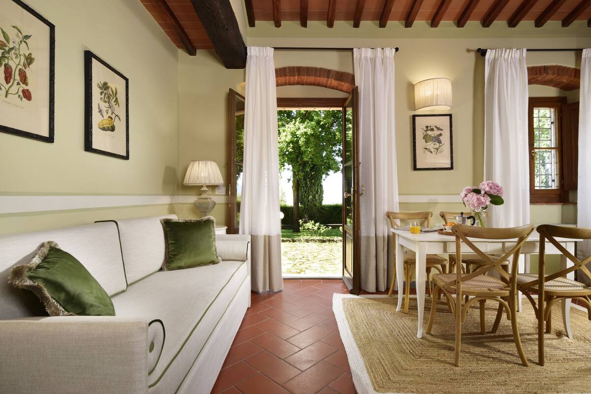 Un salon chaleureux avec un canapé blanc, des coussins verts, une table à manger en bois avec des chaises, et une vue sur un jardin par une porte ouverte.