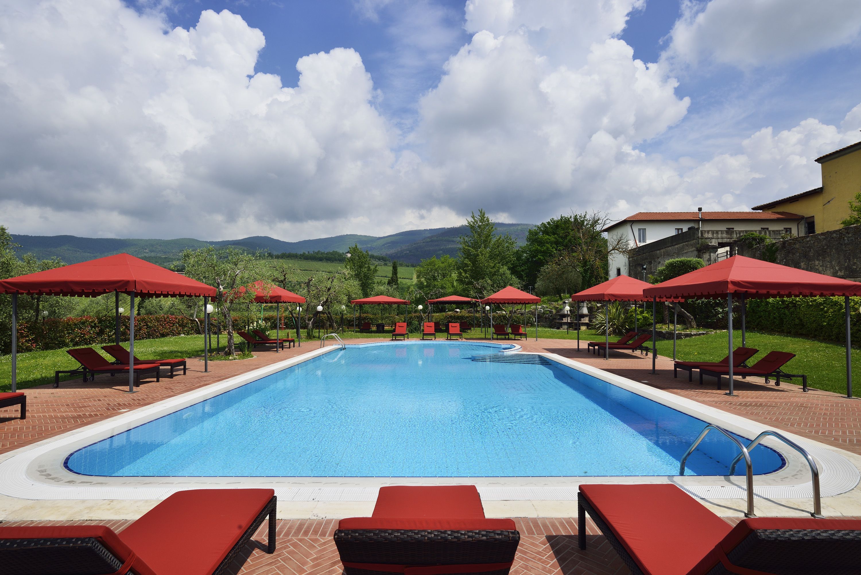 Ein Pool mit roten Sonnenschirmen und Liegestühlen, umgeben von Pflastersteinen und Grünpflanzen.