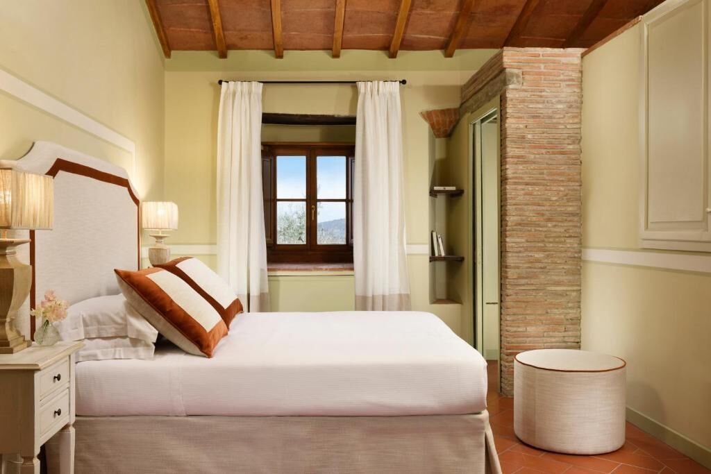 Un dormitorio con cama, mesita de noche, lámpara, ventana con cortinas, pared de ladrillo y puerta.