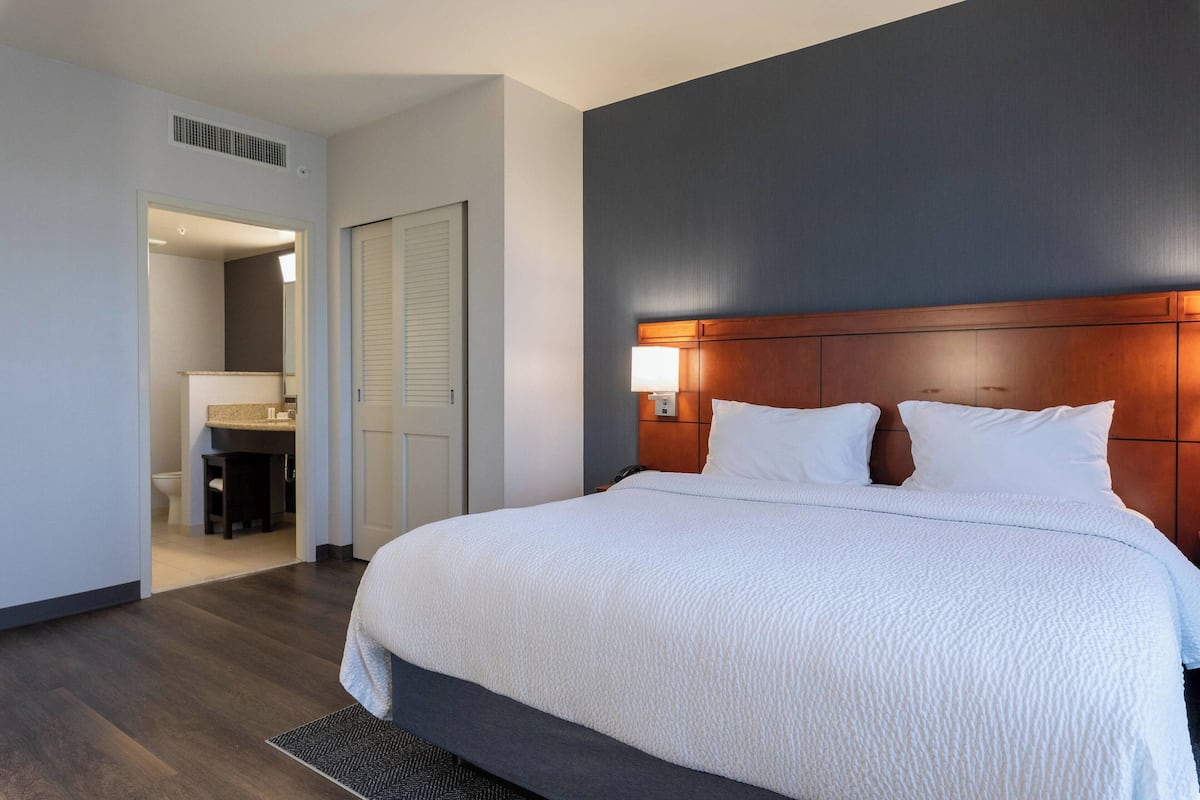 A hotel room with a large bed, a wooden headboard, a nightstand with a lamp, and a bathroom visible through an open door.