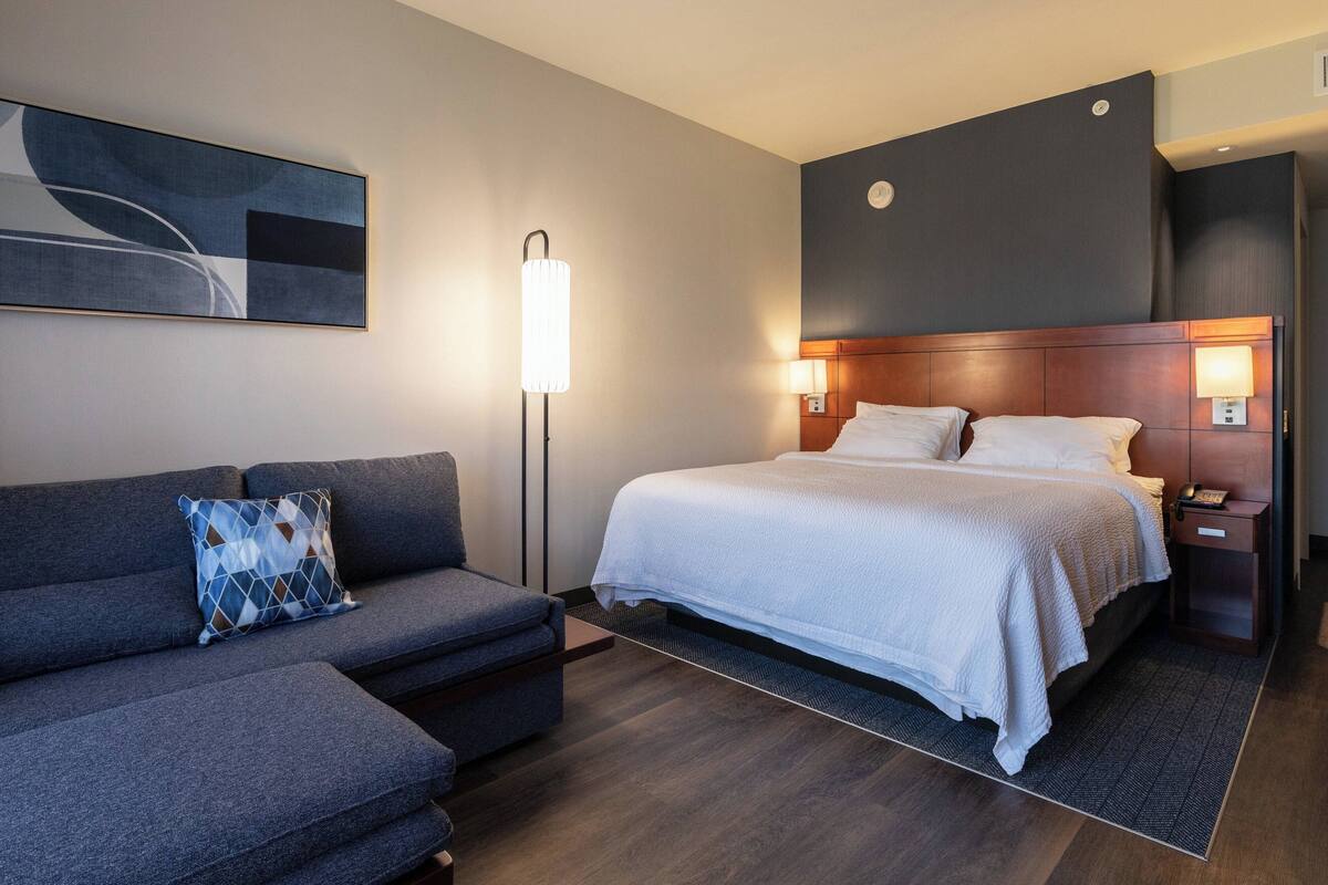 A modern hotel room with a bed, a sofa, a standing lamp, and a wall-mounted artwork.