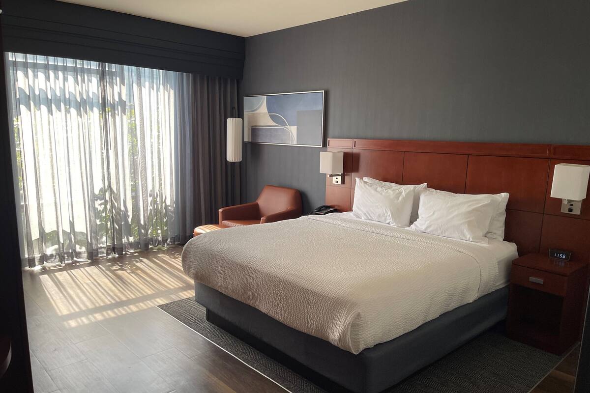 A modern hotel room with a large bed, a chair, a bedside table, and a view of the outdoors through sheer curtains.