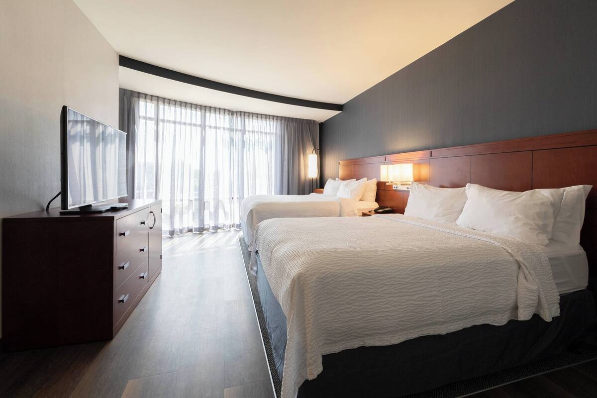 A hotel room with two beds, a TV, a dresser, and a large window with curtains.