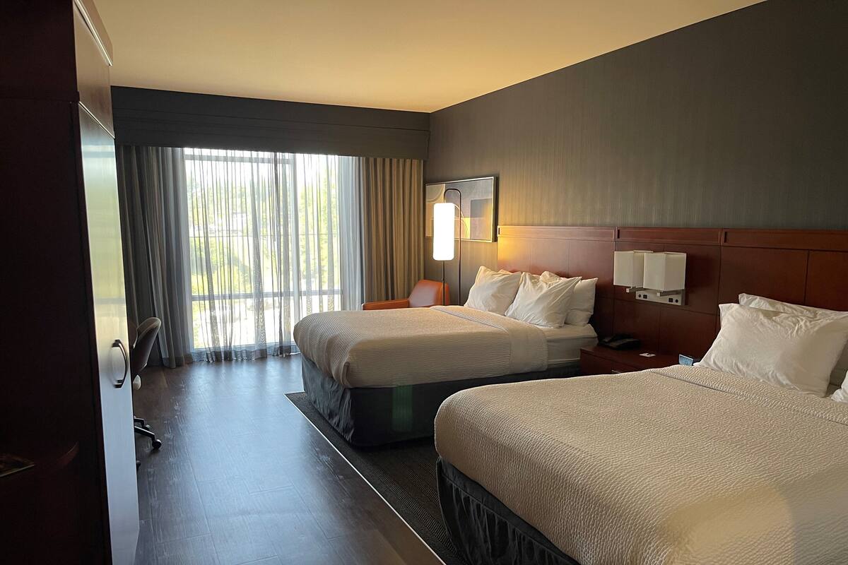 A hotel room with two beds, a large window with curtains, a bedside table with a lamp, and a wall-mounted light fixture.