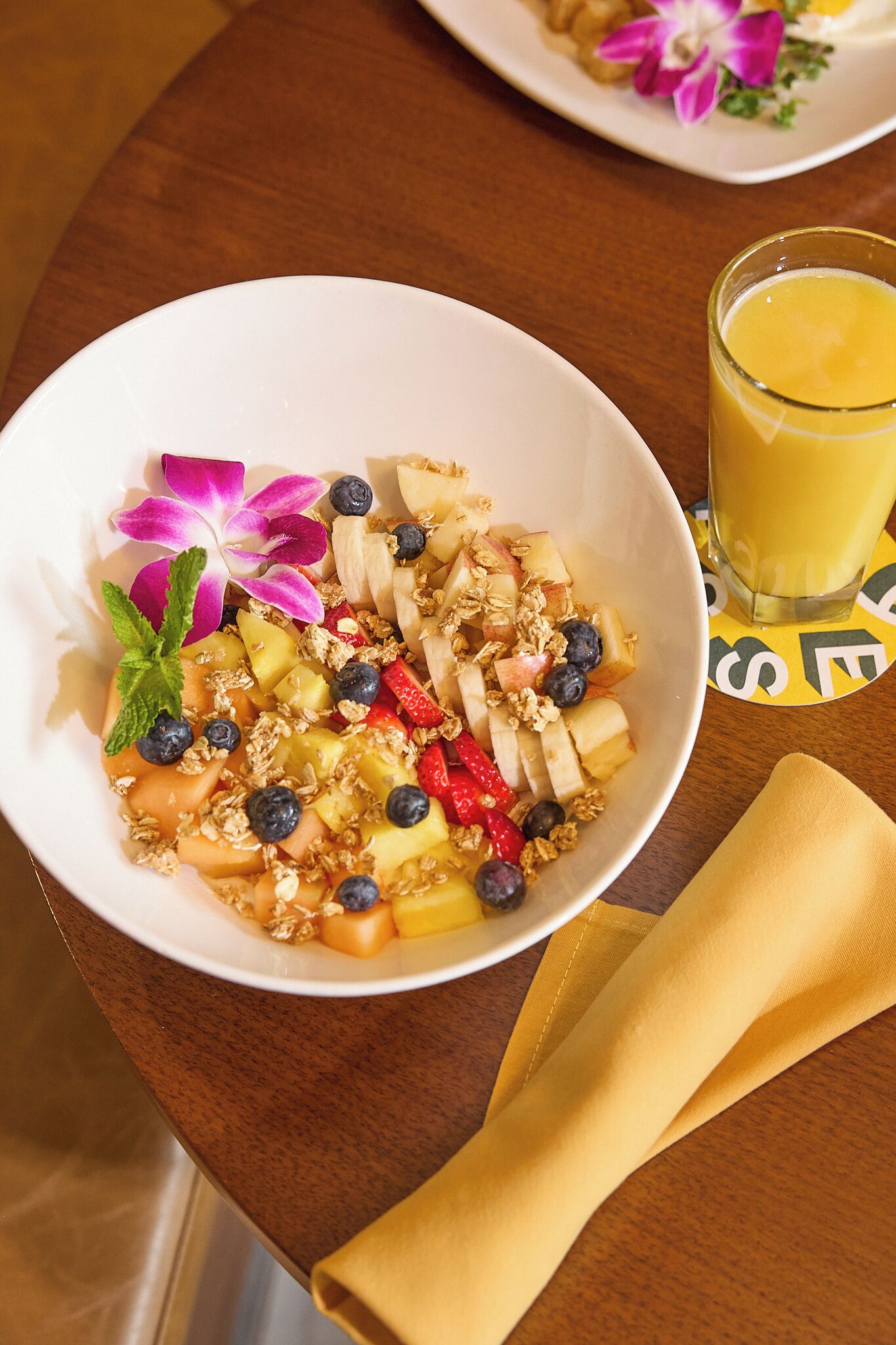 Uma saladeira com salada de frutas, granola e uma flor decorativa, acompanhada de um copo de suco de laranja sobre uma mesa de madeira.