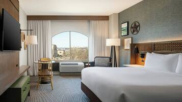 A hotel room with a large bed, a desk with a chair, a flat-screen TV, and a large window with curtains.