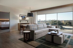 A modern hotel room with a large window, a sofa, a dining area, and a bed.